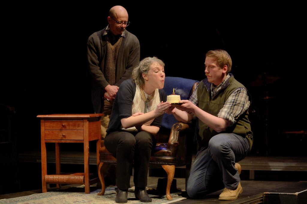 3 actors on stage. Woman blows out a birthday candle on a small cake that another actor is holding.