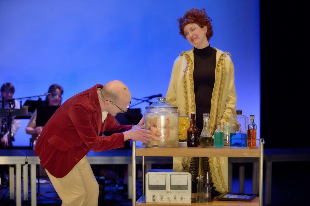 Two actors on stage. One actor is looking at a plastic head in a jar of liquid while the other actor is looking at him.