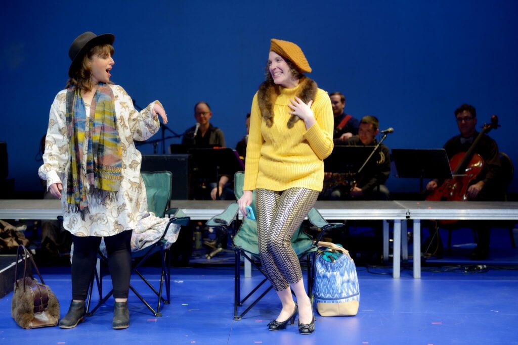 Two actors on stage in hipster costumes with a band behind them.