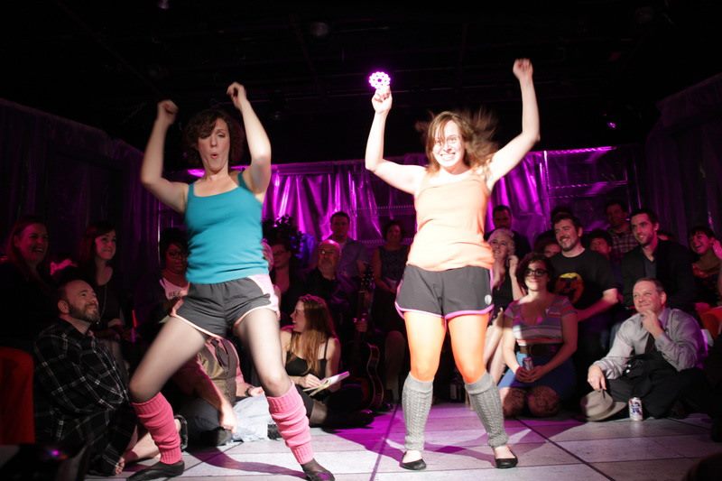 Two performers dancing on stage with two dozen people sitting behind them.