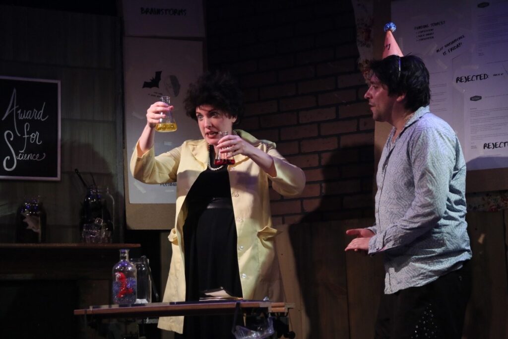 Two actors on stage. Actor on the left is a scientist looking at two beakers. Actor on the right is wearing a birthday hat.