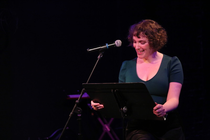 Woman with curly brown hair sitting on a stool while reading a script off of a music stand.