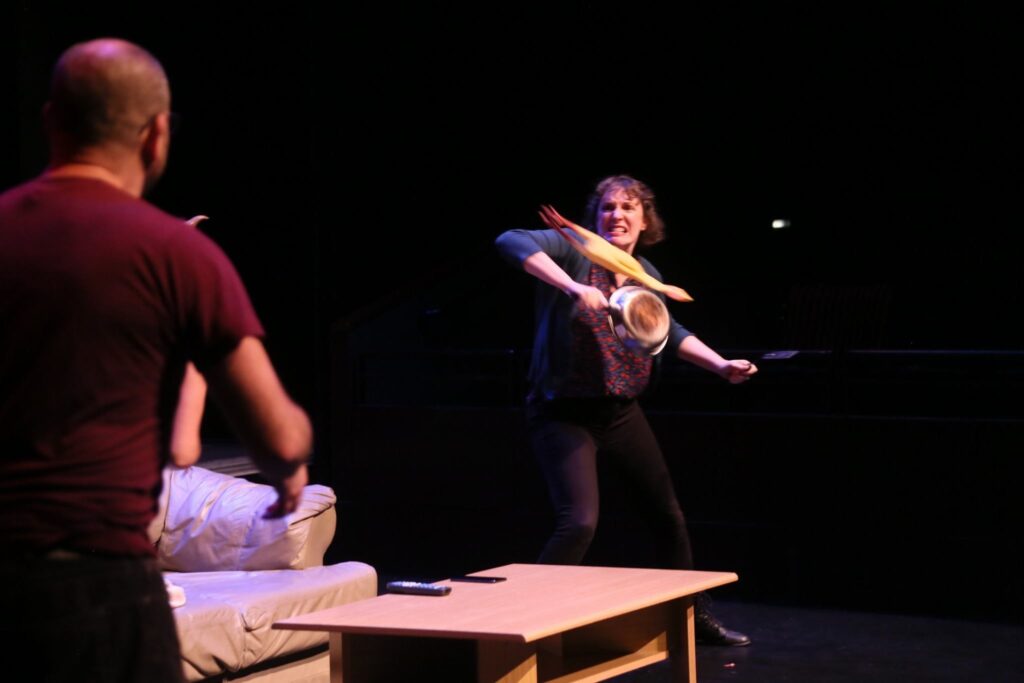 Actor on stage about to hit a rubber chicken flying towards her with a frying pan.
