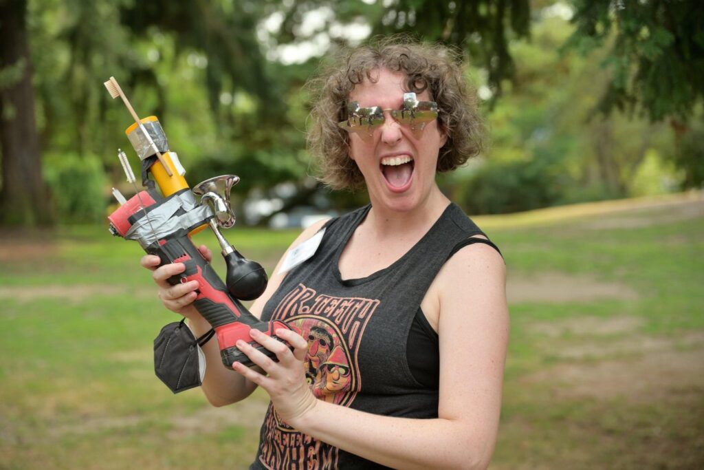 Woman wearing Texas-shaped sunglasses and holding an alarming drill contraption while in a park on a sunny day.