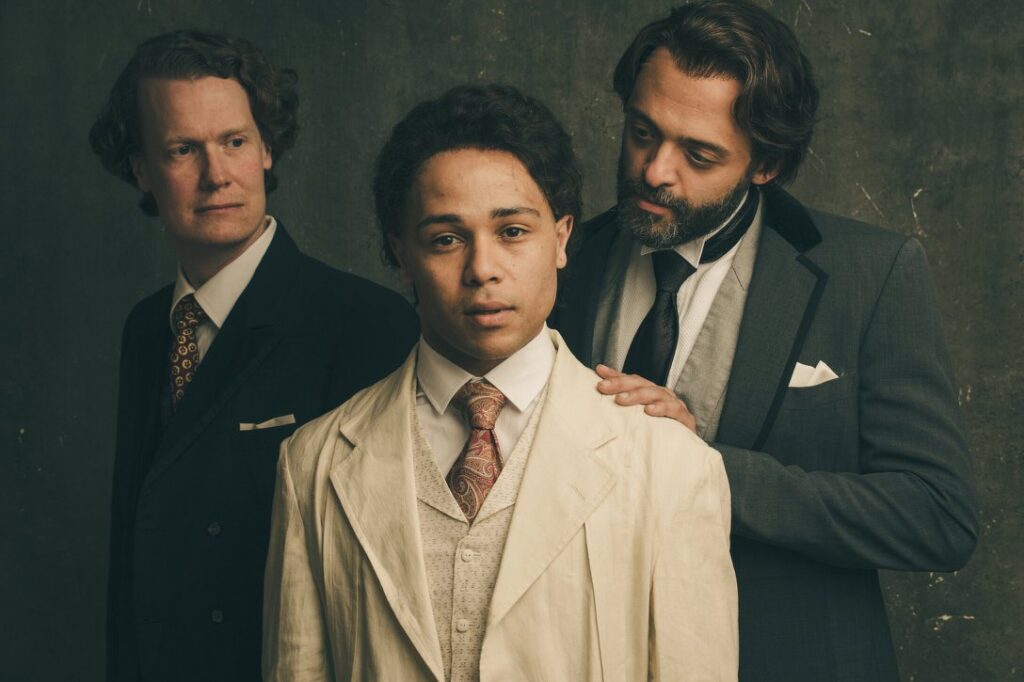 3 actors standing in front of a dark backdrop. Two actors are looking at the center actor while the center actor looks at the camera.