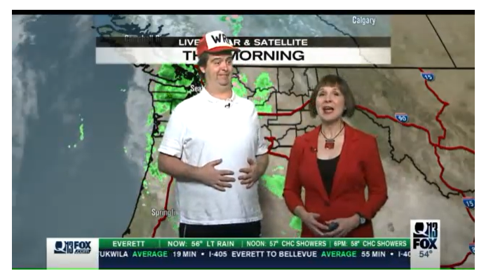 Actor dressed as Homer Simpson in front of a weather green screen. He is standing next to a meteorologist in a red blazer.