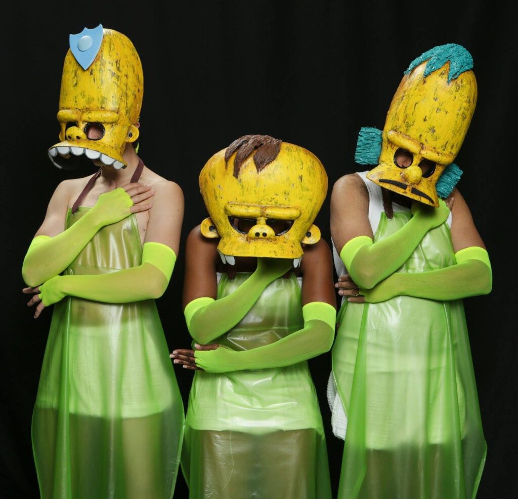 Three actors against a black backdrop. They are wearing giant masks of characters from The Simpsons.