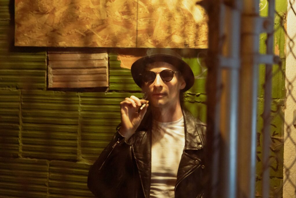 An actor behind a fence at night looking at the camera with a cigarette in his hand.