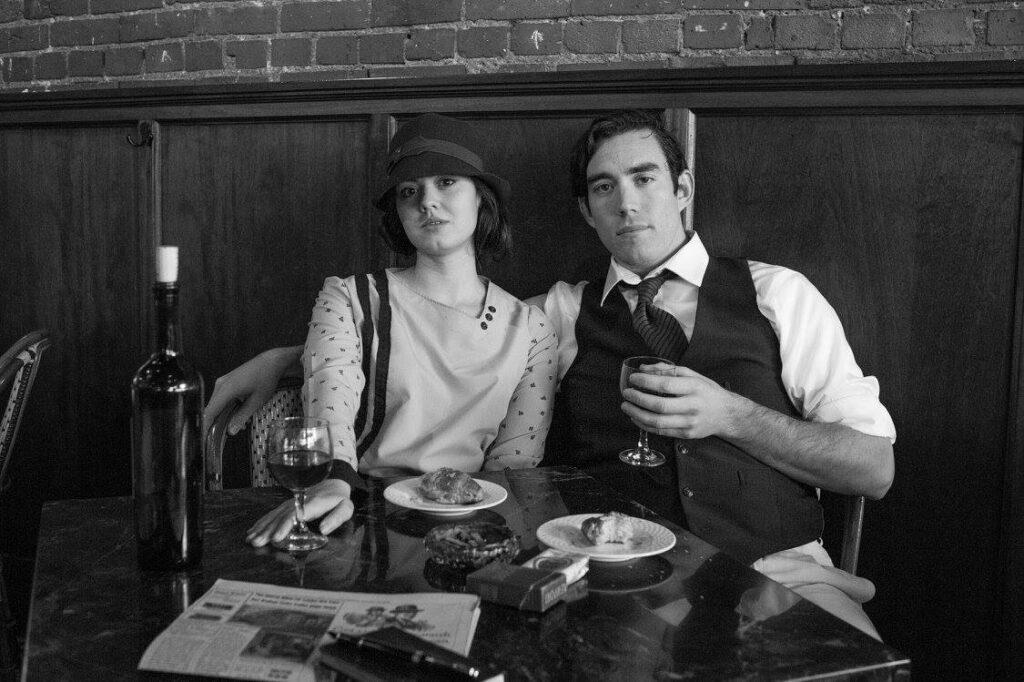 Two actors dressed as Ernest and Hadley Hemingway in a cafe. Their table has a wine bottle, wine glasses, a newspaper, cigarettes, and two small plates with one croissant on each plate.